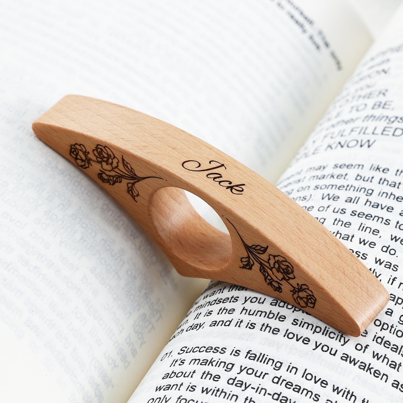 May include: A light-colored wooden thumb page holder featuring a central circular opening for a thumb. The surface is engraved with the name Jack in a cursive script and two floral rose designs on either side of the center hole.