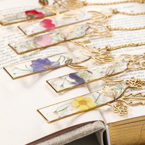 Puede incluir: Una colecci&oacute;n de marcadores hechos a mano con flores prensadas encapsuladas en resina transparente. Cada marcador tiene un marco de metal dorado y un dise&ntilde;o floral decorativo en la parte superior, con una cadena.