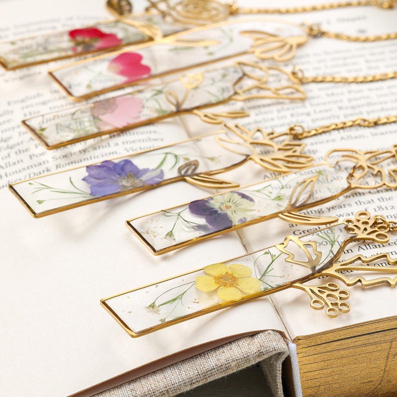 Puede incluir: Una colecci&oacute;n de marcadores hechos a mano con flores prensadas encapsuladas en resina transparente. Cada marcador tiene un marco de metal dorado y un dise&ntilde;o floral decorativo en la parte superior, con una cadena.