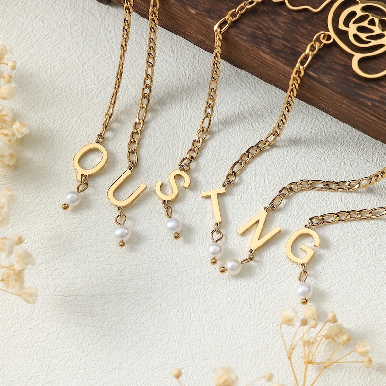 Puede incluir: Collares dorados que deletrean la palabra "QUSTNG". Cada letra es un colgante en una cadena, con un peque&ntilde;o dije de perla colgando debajo. Los collares se exhiben sobre una superficie de color claro.