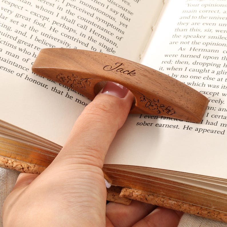 May include: A curved, brown wooden thumb page holder featuring a central circular opening for a finger. The surface is engraved with the name Jack in a cursive script and delicate floral patterns on both sides, resting on an open book page.