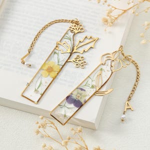 Puede incluir: Dos marcap&aacute;ginas rectangulares dorados con flores secas encapsuladas en resina transparente. Uno presenta una flor amarilla, el otro una flor morada. Ambos tienen detalles de cadena dorada y recortes florales decorativos.