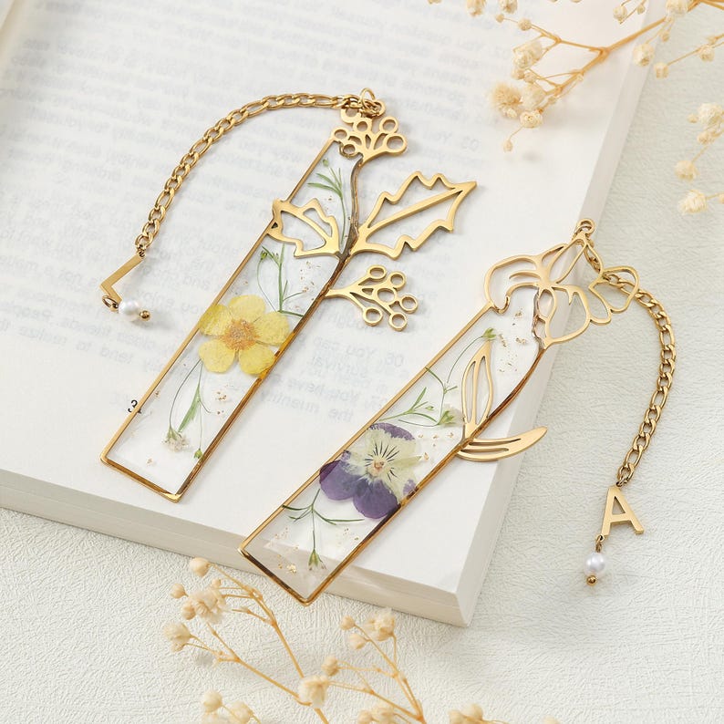 Puede incluir: Dos marcap&aacute;ginas rectangulares dorados con flores secas encapsuladas en resina transparente. Uno presenta una flor amarilla, el otro una flor morada. Ambos tienen detalles de cadena dorada y recortes florales decorativos.