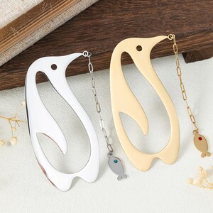 May include: Two metal penguin-shaped bookmarks, one silver-toned and one gold-toned. Each features a cutout design, a small circular hole at the top, and a delicate chain attached to a small fish charm with a single embedded crystal.