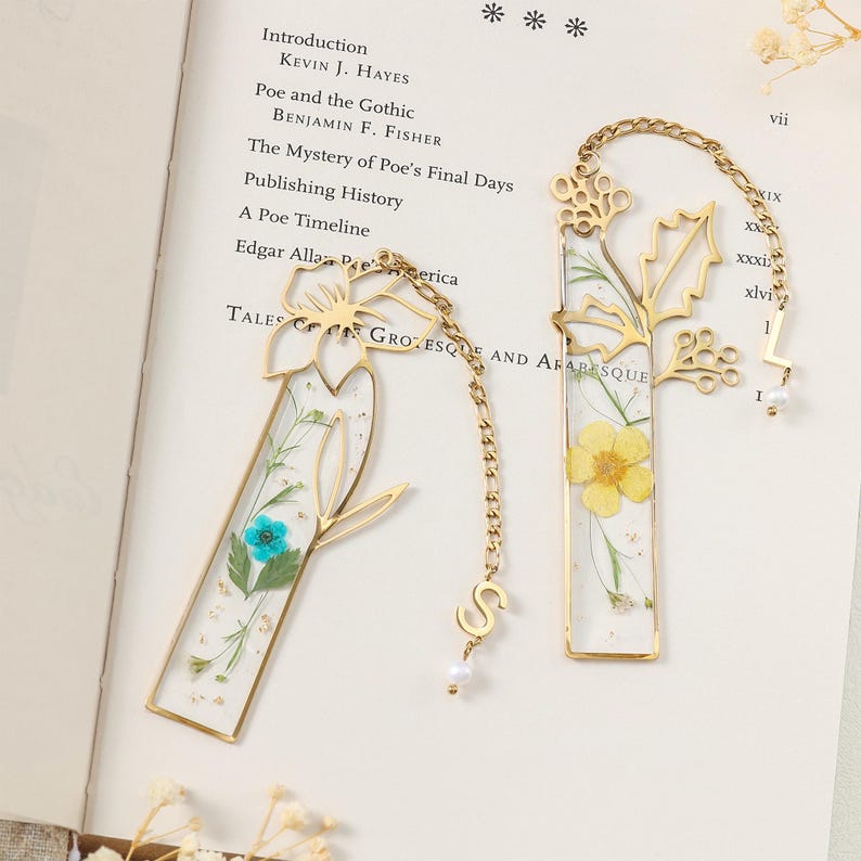 Puede incluir: Dos marcap&aacute;ginas de metal dorado con dise&ntilde;os florales. Uno presenta una flor azul, el otro una flor amarilla, ambos encapsulados en resina transparente. Cada uno tiene una cadena con un dije de letra y una peque&ntilde;a perla.