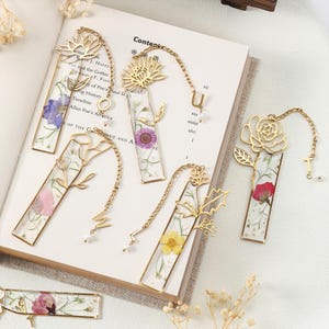 Puede incluir: Una colecci&oacute;n de marcadores de metal dorado con dise&ntilde;os florales y flores prensadas encapsuladas en resina transparente. Cada marcador presenta una flor diferente y una cadena decorativa con un dije. Los marcadores se muestran en un libro abierto.