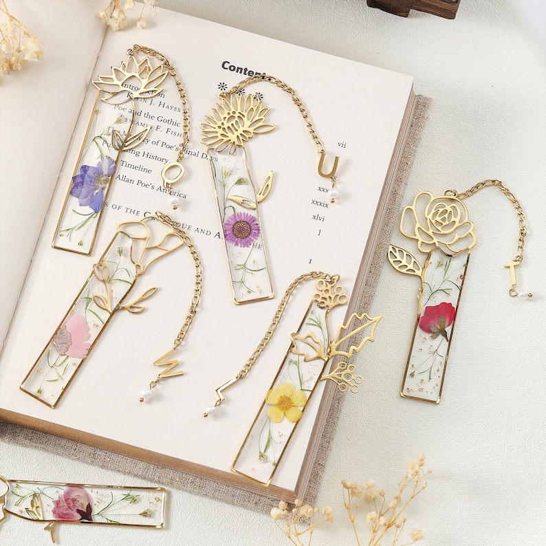 Puede incluir: Una colecci&oacute;n de marcadores de metal dorado con dise&ntilde;os florales y flores prensadas encapsuladas en resina transparente. Cada marcador presenta una flor diferente y una cadena decorativa con un dije. Los marcadores se muestran en un libro abierto.
