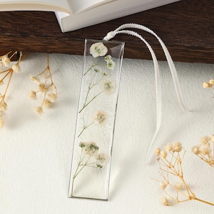 May include: A clear, rectangular bookmark featuring dried white flowers and green stems set in resin. A white ribbon is attached at the top. The bookmark rests on a white surface, with a book visible in the background.
