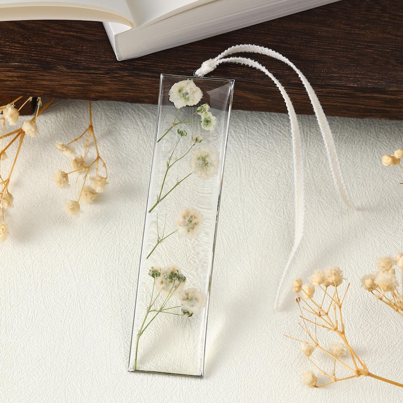 May include: A clear, rectangular bookmark featuring dried white flowers and green stems set in resin. A white ribbon is attached at the top. The bookmark rests on a white surface, with a book visible in the background.