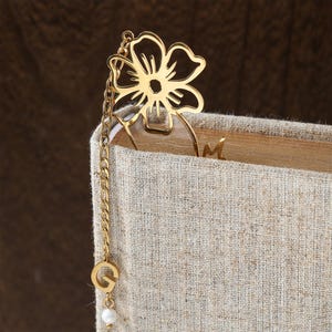 Puede incluir: Un marcap&aacute;ginas de metal dorado con un dise&ntilde;o de flor. El marcap&aacute;ginas est&aacute; unido a una cadena con un dije de la letra "G" y una peque&ntilde;a perla blanca. El marcap&aacute;ginas est&aacute; insertado en un libro con una cubierta de lino beige.
