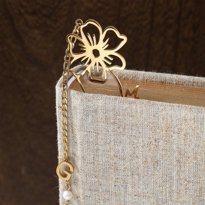 Puede incluir: Un marcap&aacute;ginas de metal dorado con un dise&ntilde;o de flor. El marcap&aacute;ginas est&aacute; unido a una cadena con un dije de la letra "G" y una peque&ntilde;a perla blanca. El marcap&aacute;ginas est&aacute; insertado en un libro con una cubierta de lino beige.