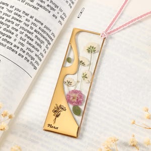 Può includere: Un segnalibro color oro con un design floreale e il nome "Nora" inciso. Il segnalibro presenta fiori pressati in resina trasparente e un cordoncino rosa e bianco. Il segnalibro è rettangolare con un ritaglio decorativo.