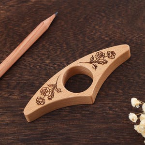 May include: A light brown wooden tatting shuttle featuring a central circular opening and a delicate dark brown laser-engraved rose floral pattern on both tapered ends, positioned next to a wooden pencil on a dark brown textured surface.