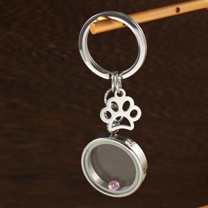 May include: Silver-coloured keyring with a paw print charm and a round locket containing a pink bead. The keyring hangs from a gold-coloured rod against a dark brown backdrop. A simple and elegant accessory.