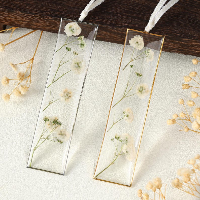 May include: Two rectangular clear resin bookmarks, one with a silver border and the other with a gold border. Each bookmark features delicate white flowers and green stems. White ribbon is attached at the top for easy use.