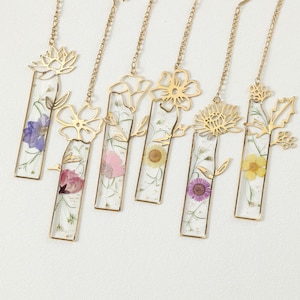 Puede incluir: Colecci&oacute;n de seis marcadores de metal dorado con dise&ntilde;os florales. Cada marcador presenta una secci&oacute;n rectangular de resina transparente con una flor prensada y una cadena dorada. Las flores son de varios colores, incluyendo morado, rosa y amarillo.