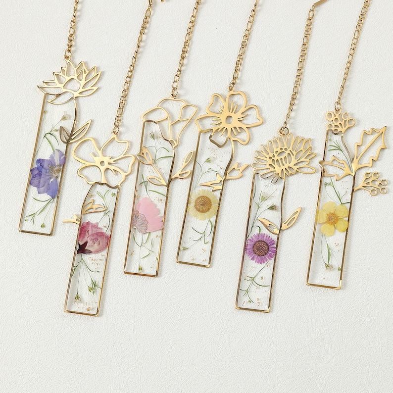 Puede incluir: Colecci&oacute;n de seis marcadores de metal dorado con dise&ntilde;os florales. Cada marcador presenta una secci&oacute;n rectangular de resina transparente con una flor prensada y una cadena dorada. Las flores son de varios colores, incluyendo morado, rosa y amarillo.