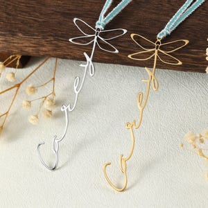 May include: Two dragonfly-shaped bookmarks. One is silver-colored, the other gold-colored. Each has a dragonfly design at the top and a cursive word design that ends in a hook. The bookmarks are attached to a teal and white ribbon.