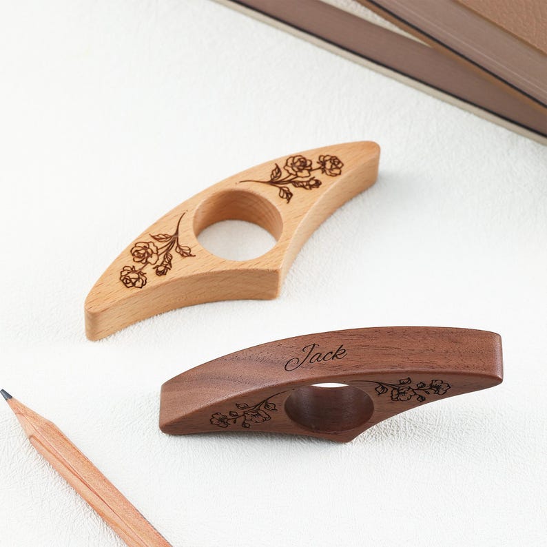 May include: Two wooden book page holders with a central hole for a thumb. The lighter wood holder features an engraved rose design. The darker wood holder features an engraved rose design and the name Jack in a cursive script font.