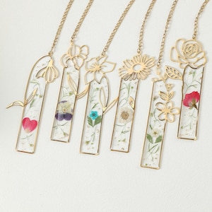 Puede incluir: Una colecci&oacute;n de seis marcap&aacute;ginas de metal dorado, cada uno con un dise&ntilde;o floral diferente. Los marcap&aacute;ginas tienen inserciones de resina transparente con flores prensadas en varios colores, incluyendo rojo, morado y azul. Cada marcap&aacute;ginas est&aacute; unido a una delicada cadena dorada.