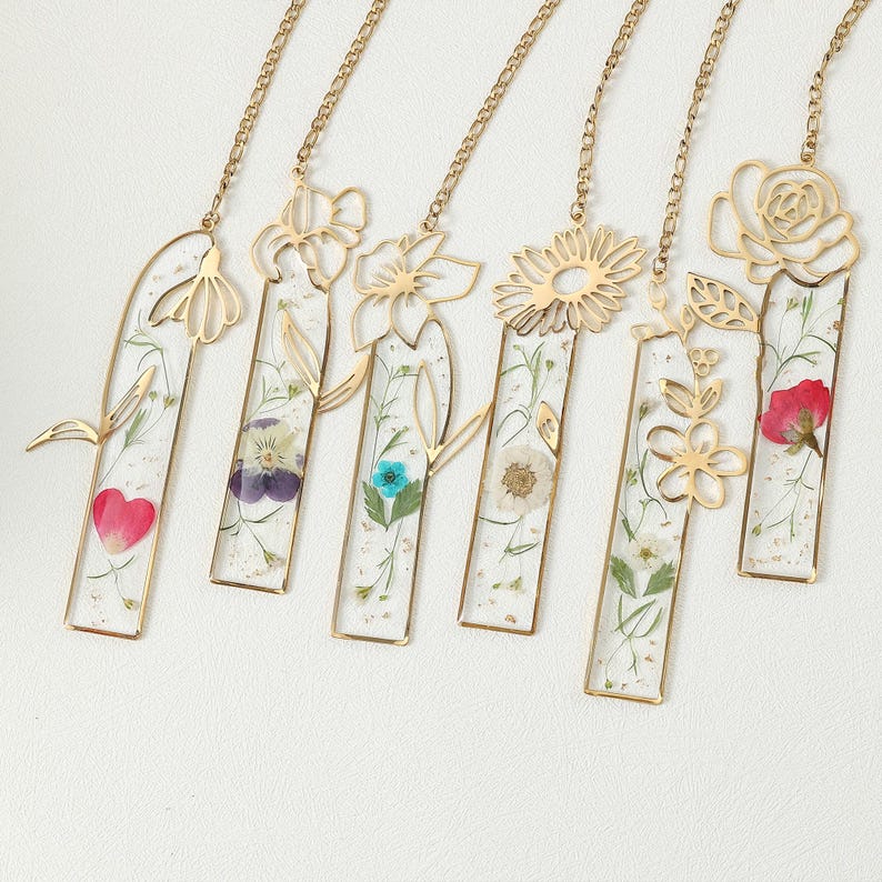 Puede incluir: Una colecci&oacute;n de seis marcap&aacute;ginas de metal dorado, cada uno con un dise&ntilde;o floral diferente. Los marcap&aacute;ginas tienen inserciones de resina transparente con flores prensadas en varios colores, incluyendo rojo, morado y azul. Cada marcap&aacute;ginas est&aacute; unido a una delicada cadena dorada.
