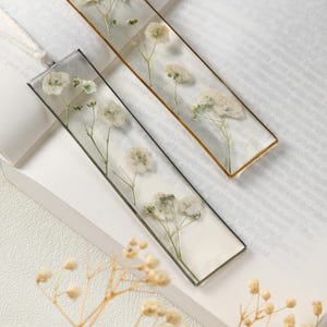 May include: Two rectangular bookmarks with dried white flowers encased in clear resin. One has a gold border, the other a black border. Both have a white ribbon attached for use.