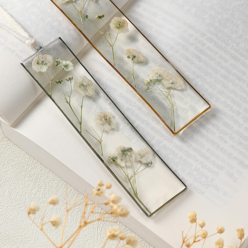 May include: Two rectangular bookmarks with dried white flowers encased in clear resin. One has a gold border, the other a black border. Both have a white ribbon attached for use.