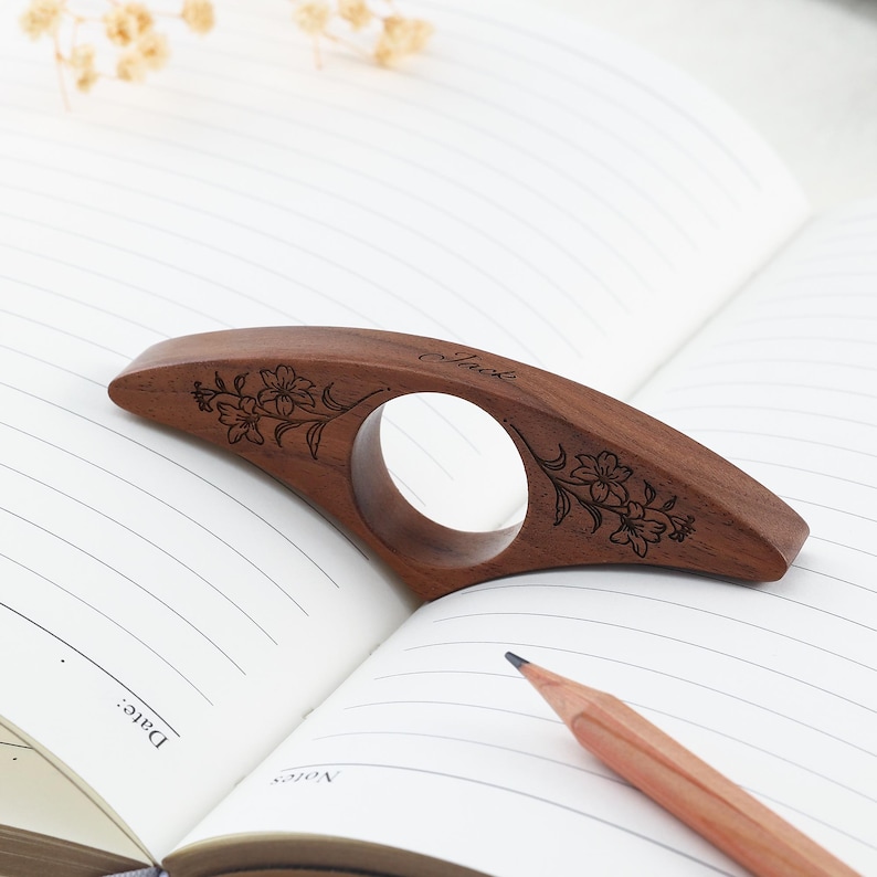 May include: A curved, dark brown wooden book page holder with a central circular opening. The wood features an engraved floral pattern on both sides and the name Jack inscribed in cursive script on the top edge of the curved wooden surface.