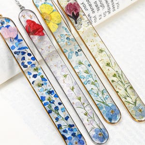 May include: A collection of five handcrafted resin bookmarks. Each bookmark features a unique design with embedded dried flowers, blue and gold accents, and a gold-colored metal border. The bookmarks are arranged diagonally, showcasing their individual floral arrangements.