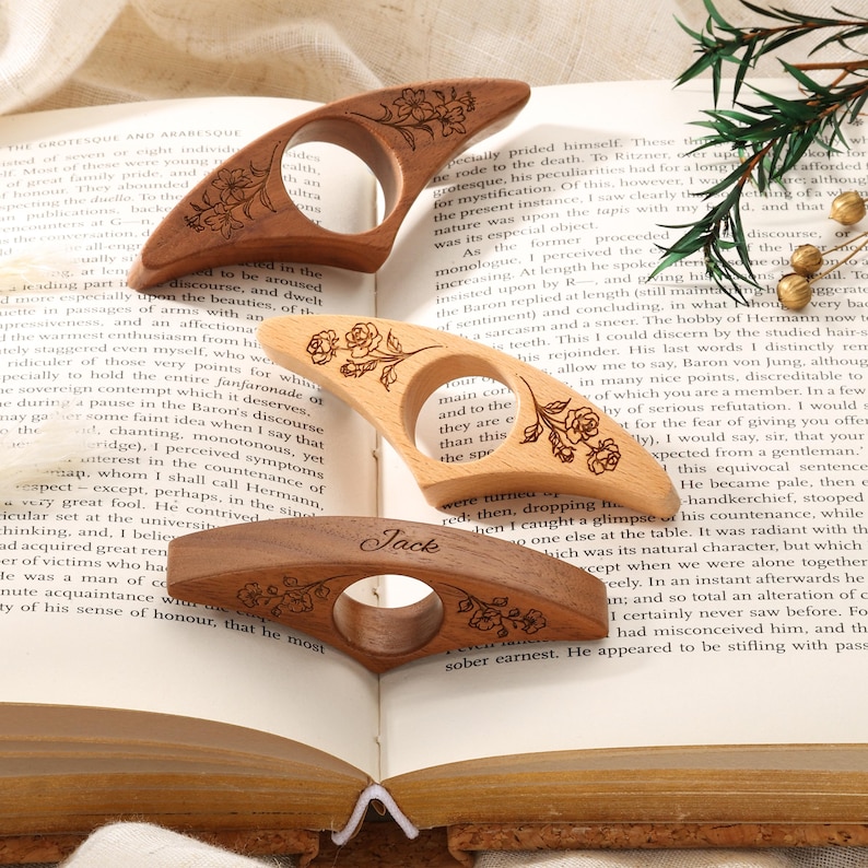May include: Three wooden page holders with a central hole for a thumb. The top one is dark wood with floral engravings, the middle is light wood with rose engravings, and the bottom is medium wood with floral engravings and the name Jack.