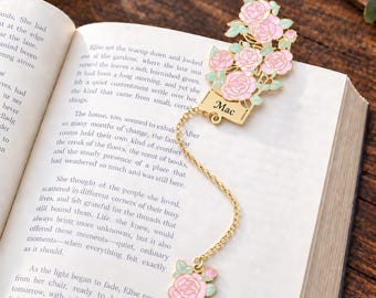 May include: A gold-toned metal bookmark featuring a cluster of pink enamel roses with light green leaves. A rectangular tag engraved with the name Mac is attached to a gold chain, which connects to a smaller matching pink rose charm at the end.
