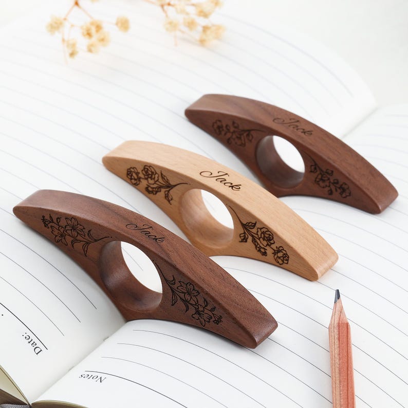 May include: Three wooden page holders with a central circular opening, each featuring a laser-engraved floral design and the name Jack. Two holders are made of dark brown wood, and one holder is made of a lighter, natural-toned wood.
