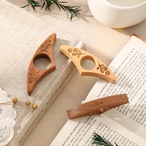 May include: Three wooden page holders with a central hole for a thumb. Two holders feature engraved floral designs, while the third is engraved with the name Jack. These accessories are crafted from light and medium-toned wood with smooth finishes.