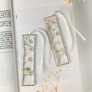 May include: Two rectangular bookmarks featuring dried white flowers set in clear resin. One has a gold frame, the other a black frame. Each has a white ribbon attached for use as a bookmark.