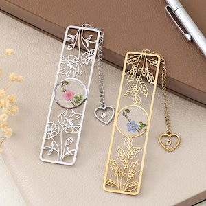 May include: Two decorative metal bookmarks. One is silver with a floral design and a pink flower encased in resin. The other is gold with a holly design and a blue flower encased in resin. Both have chain tassels with heart charms.