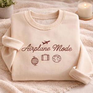May include: A cream-colored sweatshirt with the embroidered text "Airplane Mode" and travel-themed icons. The design includes an airplane, a globe, a suitcase, and a world map. The sweatshirt has a crew neck and long sleeves.