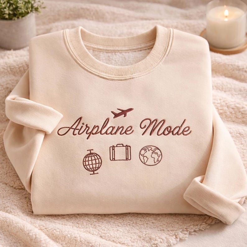 May include: A cream-colored sweatshirt with the embroidered text "Airplane Mode" and travel-themed icons. The design includes an airplane, a globe, a suitcase, and a world map. The sweatshirt has a crew neck and long sleeves.