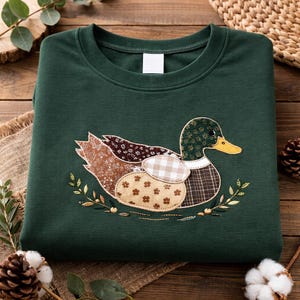 Könnte beinhalten: Ein dunkelgrünes Sweatshirt mit einer detaillierten Applikation einer Ente. Die Ente hat einen gelben Schnabel, einen grünen gemusterten Kopf und Flügel- und Körperteile aus verschiedenen gemusterten Stoffen. Das Design ist von gestickten Blättern umgeben.