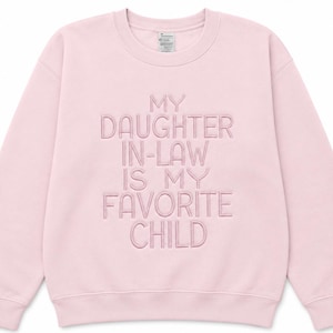 K&ouml;nnte beinhalten: Hellrosa Sweatshirt mit dem gestickten Text "MY DAUGHTER IN-LAW IS MY FAVORITE CHILD". Das Sweatshirt mit Rundhalsausschnitt hat lange &Auml;rmel und ein Champion-Etikett.