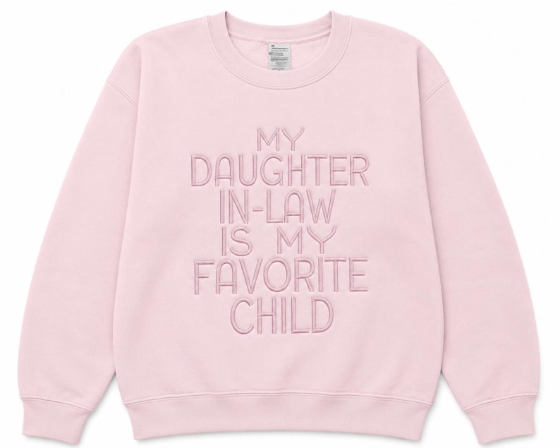 K&ouml;nnte beinhalten: Hellrosa Sweatshirt mit dem gestickten Text "MY DAUGHTER IN-LAW IS MY FAVORITE CHILD". Das Sweatshirt mit Rundhalsausschnitt hat lange &Auml;rmel und ein Champion-Etikett.