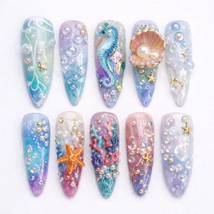 May include: A collection of ten long, almond-shaped artificial nails with intricate ocean-themed designs. Each nail features a unique combination of colors, including blues, purples, and greens, adorned with pearls, starfish, seahorses, and coral.