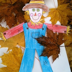 May include: A hand-drawn paper craft of a scarecrow with a brown hat, red shirt, blue overalls, and a straw-like beard. The scarecrow is surrounded by yellow and brown autumn leaves.