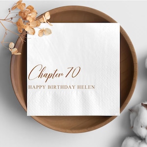 May include: A white square napkin with the text "Chapter 70 HAPPY BIRTHDAY HELEN" in a brown script font. The napkin is placed on a wooden bowl, with dried flowers and cotton accents.