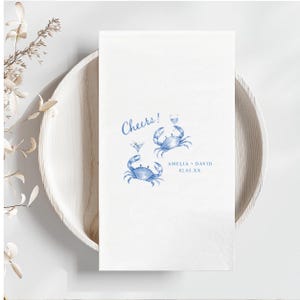 Puede incluir: Una servilleta de papel rectangular blanca con una ilustraci&oacute;n en acuarela azul de dos cangrejos sosteniendo copas de c&oacute;ctel. El texto Cheers! est&aacute; impreso en azul sobre los cangrejos, con AMELIA + DAVID y 02.03.XX impresos debajo.