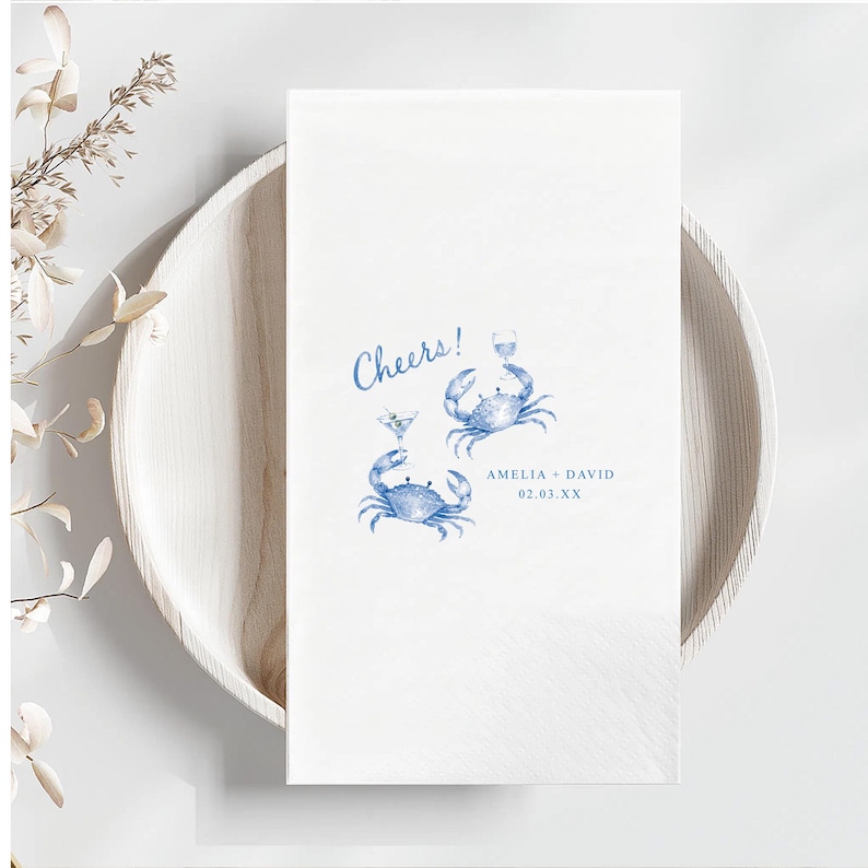 Puede incluir: Una servilleta de papel rectangular blanca con una ilustraci&oacute;n en acuarela azul de dos cangrejos sosteniendo copas de c&oacute;ctel. El texto Cheers! est&aacute; impreso en azul sobre los cangrejos, con AMELIA + DAVID y 02.03.XX impresos debajo.