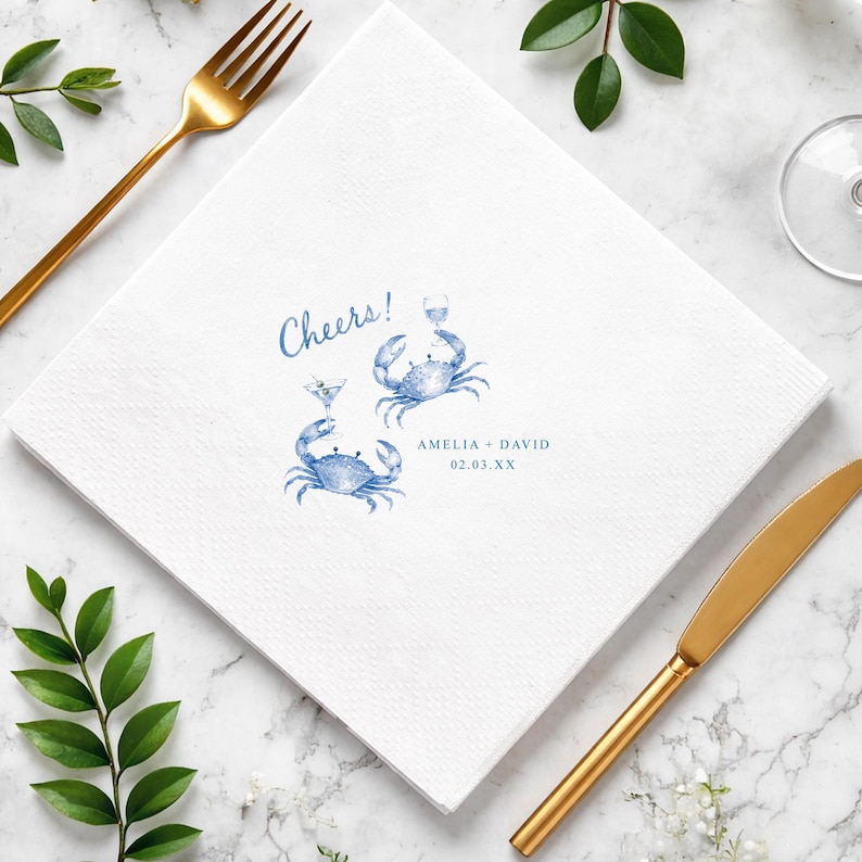 Puede incluir: Una servilleta de papel cuadrada de color blanco con una ilustraci&oacute;n en acuarela azul de dos cangrejos sosteniendo copas de martini. El texto Cheers! aparece en azul sobre los cangrejos, junto a los nombres Amelia + David y la fecha 02.03.XX.