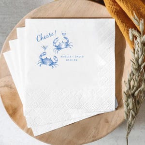 Puede incluir: Una pila de servilletas de papel blanco con un dise&ntilde;o impreso en azul de dos cangrejos sosteniendo copas de c&oacute;ctel. El texto Cheers! aparece sobre los cangrejos, con los nombres Amelia + David y la fecha 02.03.XX impresos debajo.