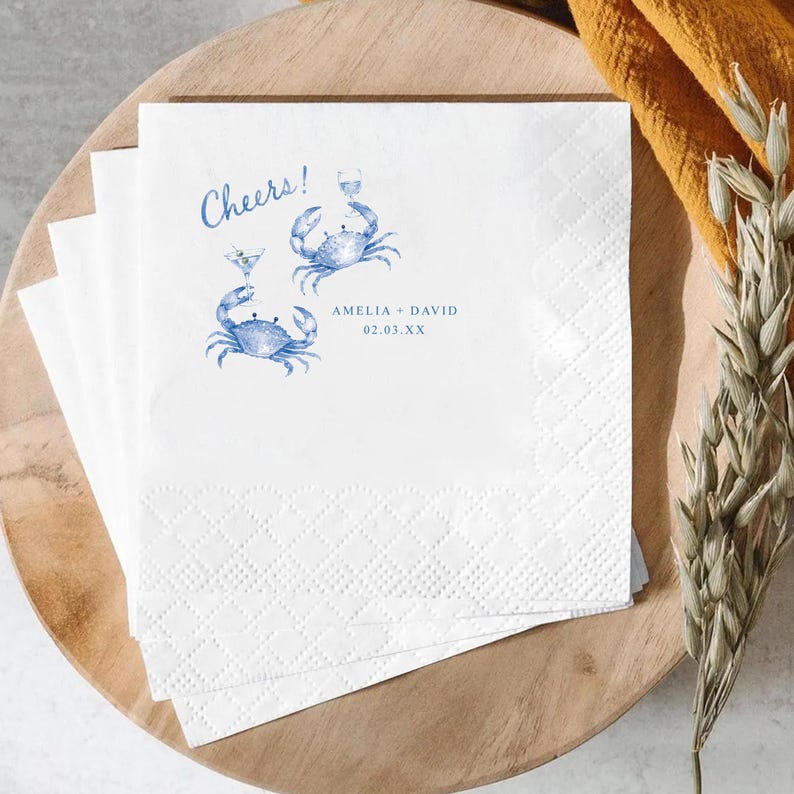 Puede incluir: Una pila de servilletas de papel blanco con un dise&ntilde;o impreso en azul de dos cangrejos sosteniendo copas de c&oacute;ctel. El texto Cheers! aparece sobre los cangrejos, con los nombres Amelia + David y la fecha 02.03.XX impresos debajo.