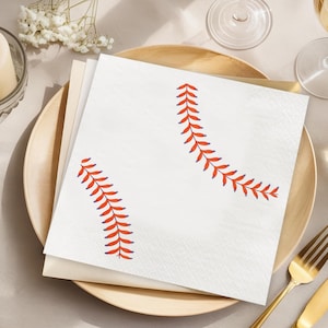 May include: White square paper napkins with a baseball stitch design in red and blue. The napkins are stacked on a wooden plate, with a fork and knife visible. The napkins are perfect for a baseball-themed party.