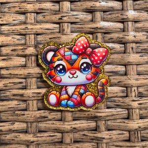 May include: A colorful, glitter-edged tiger-shaped charm with a red polka-dot bow. The tiger has large blue eyes, pink cheeks, and a patchwork design in orange, red, blue, and yellow. The charm is set against a woven background.